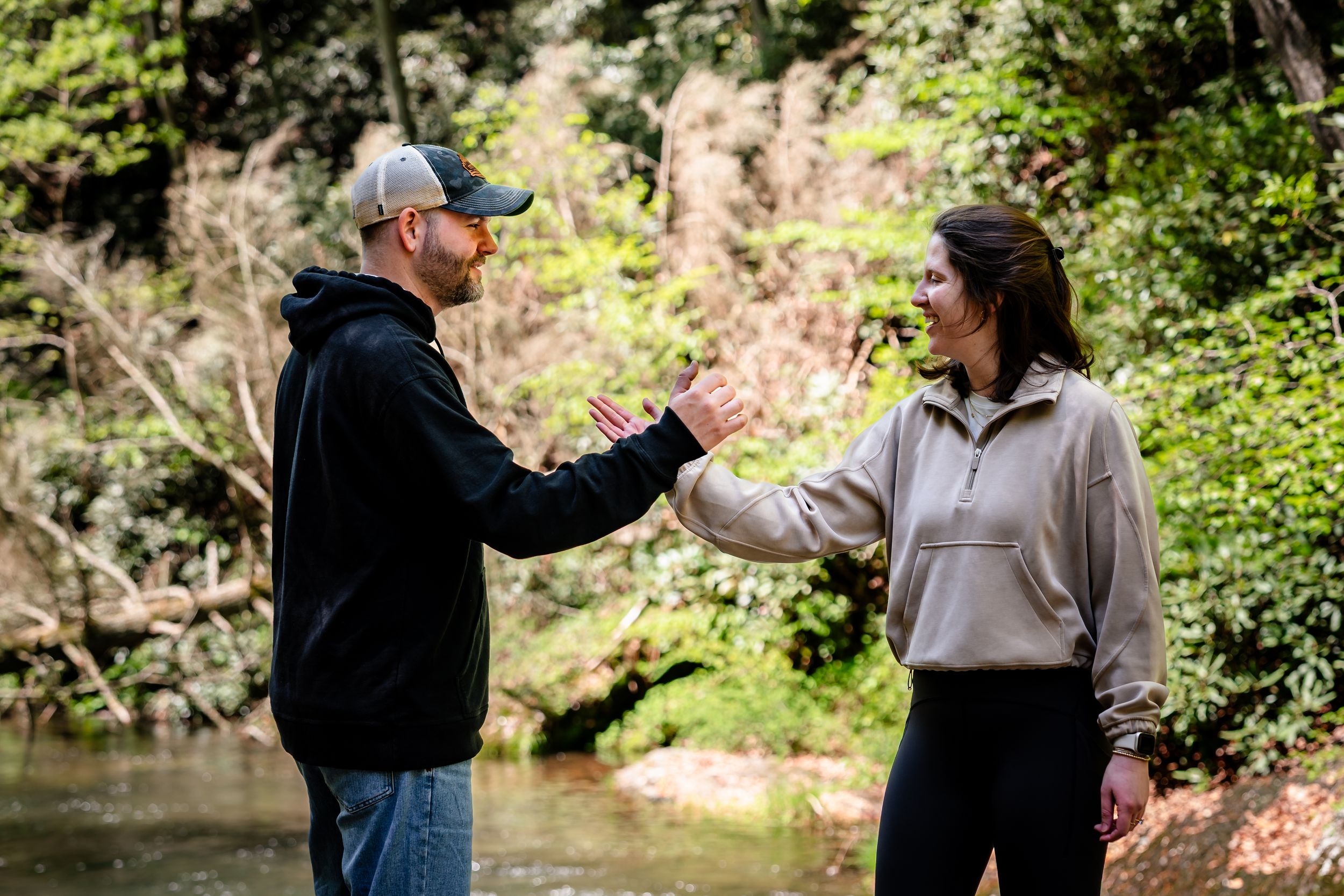 Two people interact in a sunny forest setting near water.