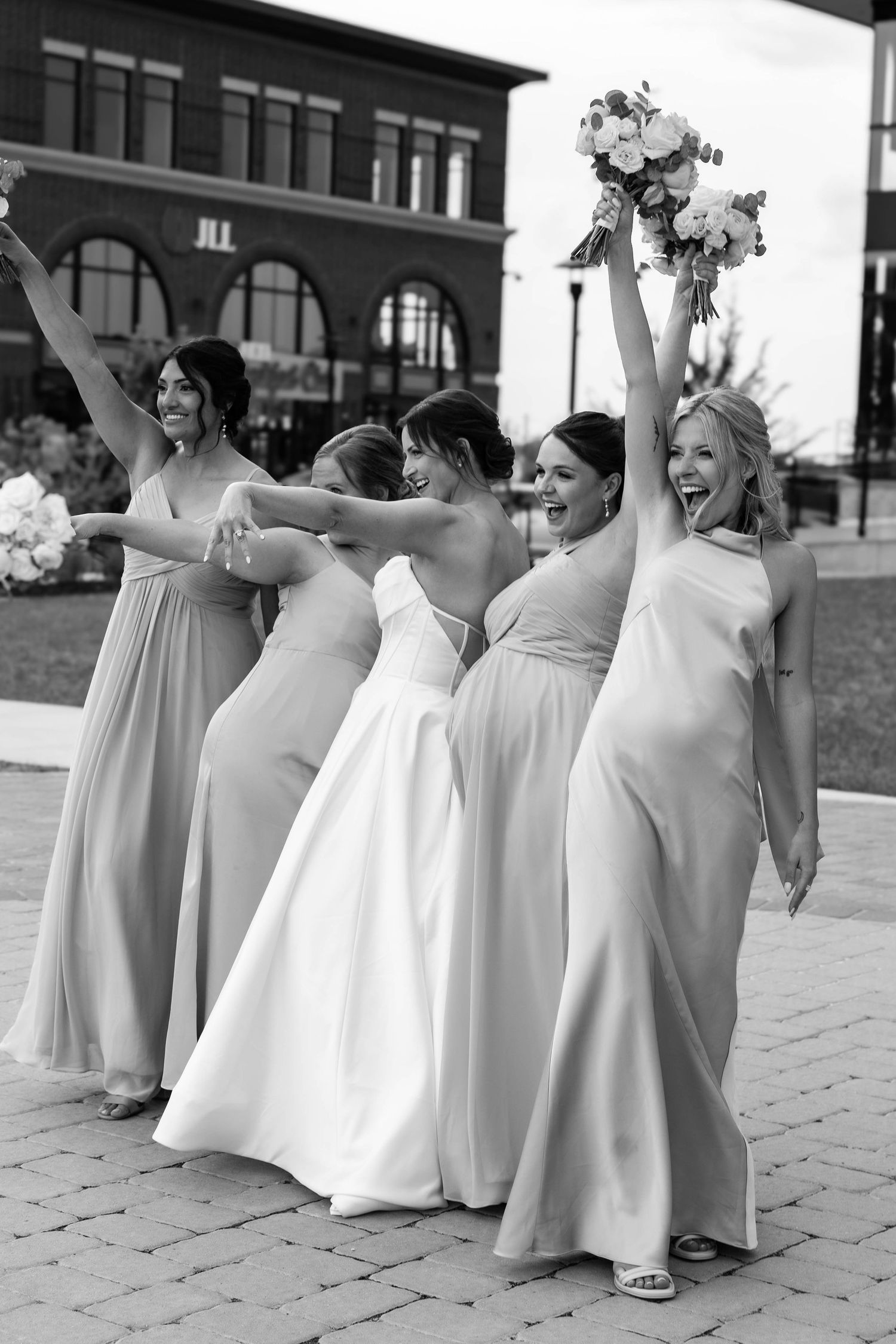 Black and white photo of a bride and bridesmaids in long gowns celebrating outdoors, raising bouquets joyfully.