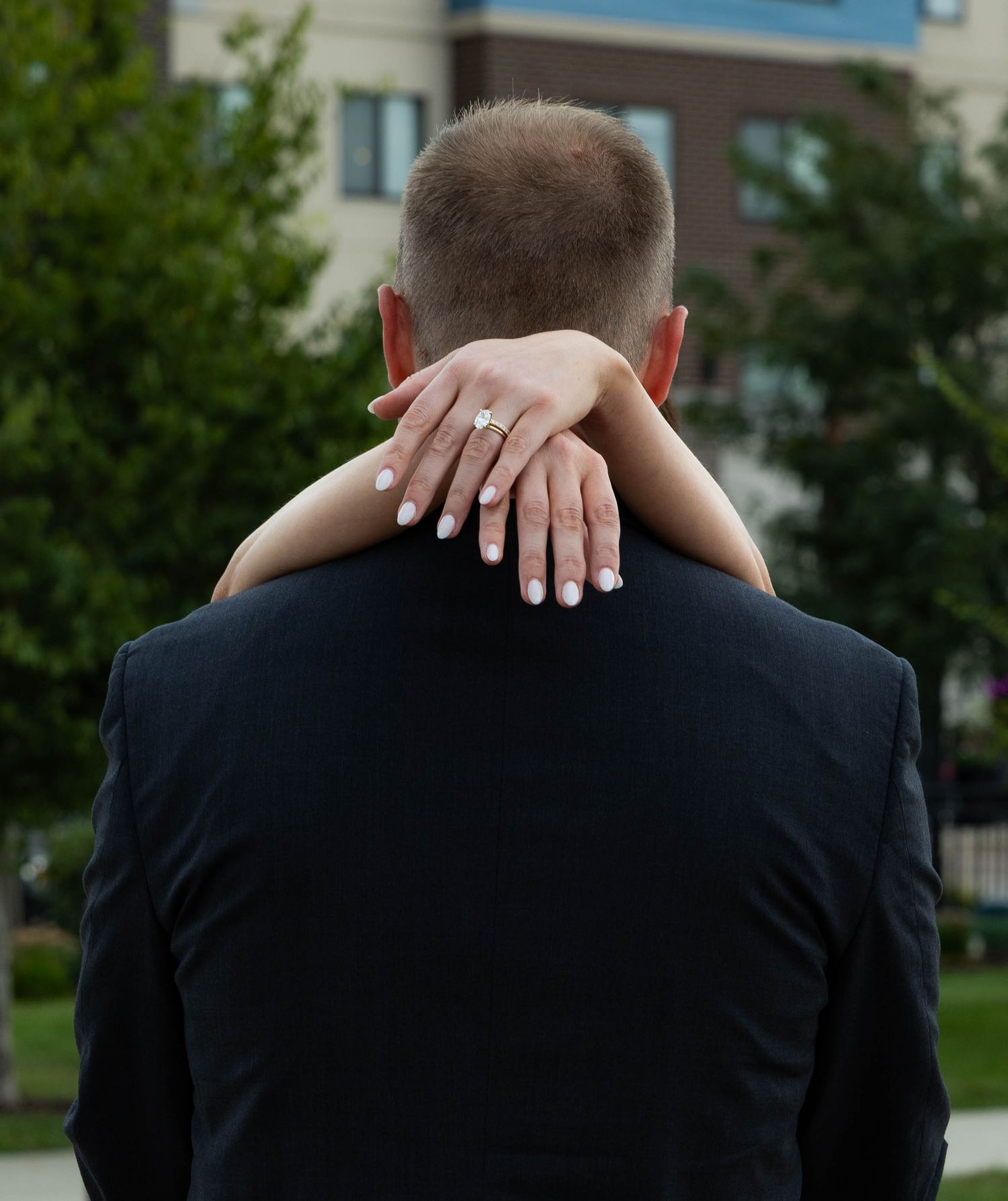 Couple embracing outdoors, woman's hand showing diamond engagement ring resting on man's shoulder in black suit.