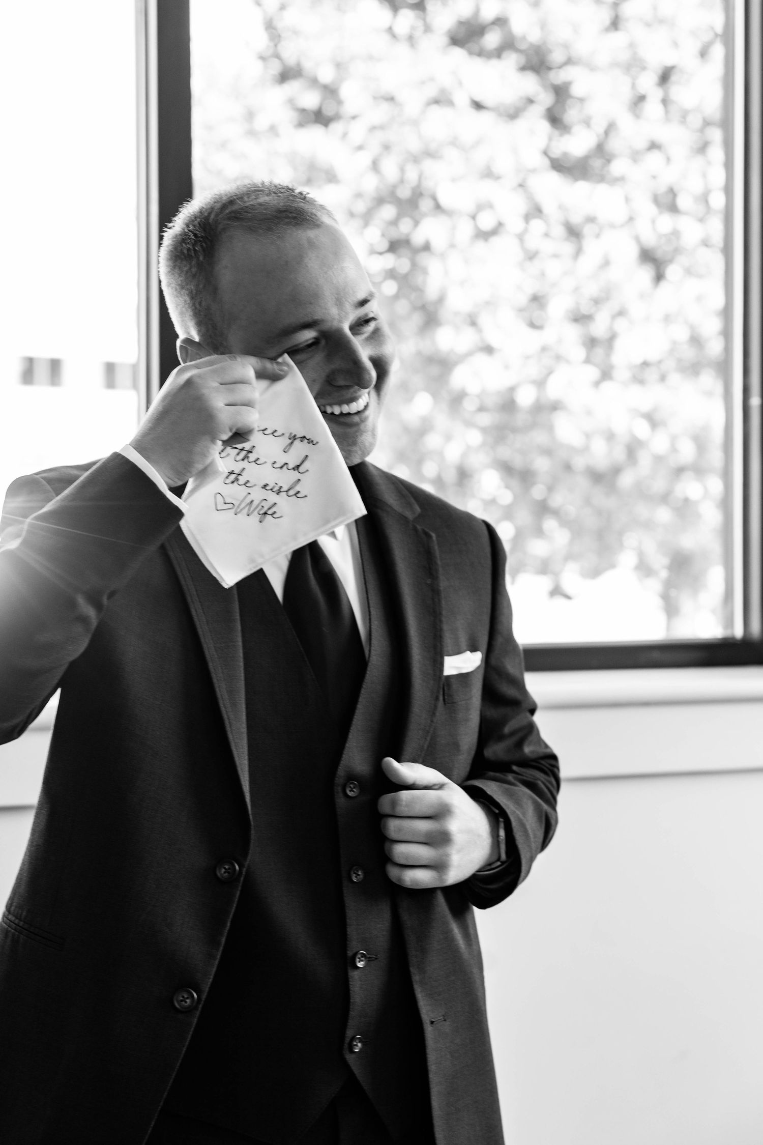 A groom in a black suit laughs while reading a handwritten note, captured in an elegant black and white wedding photo.