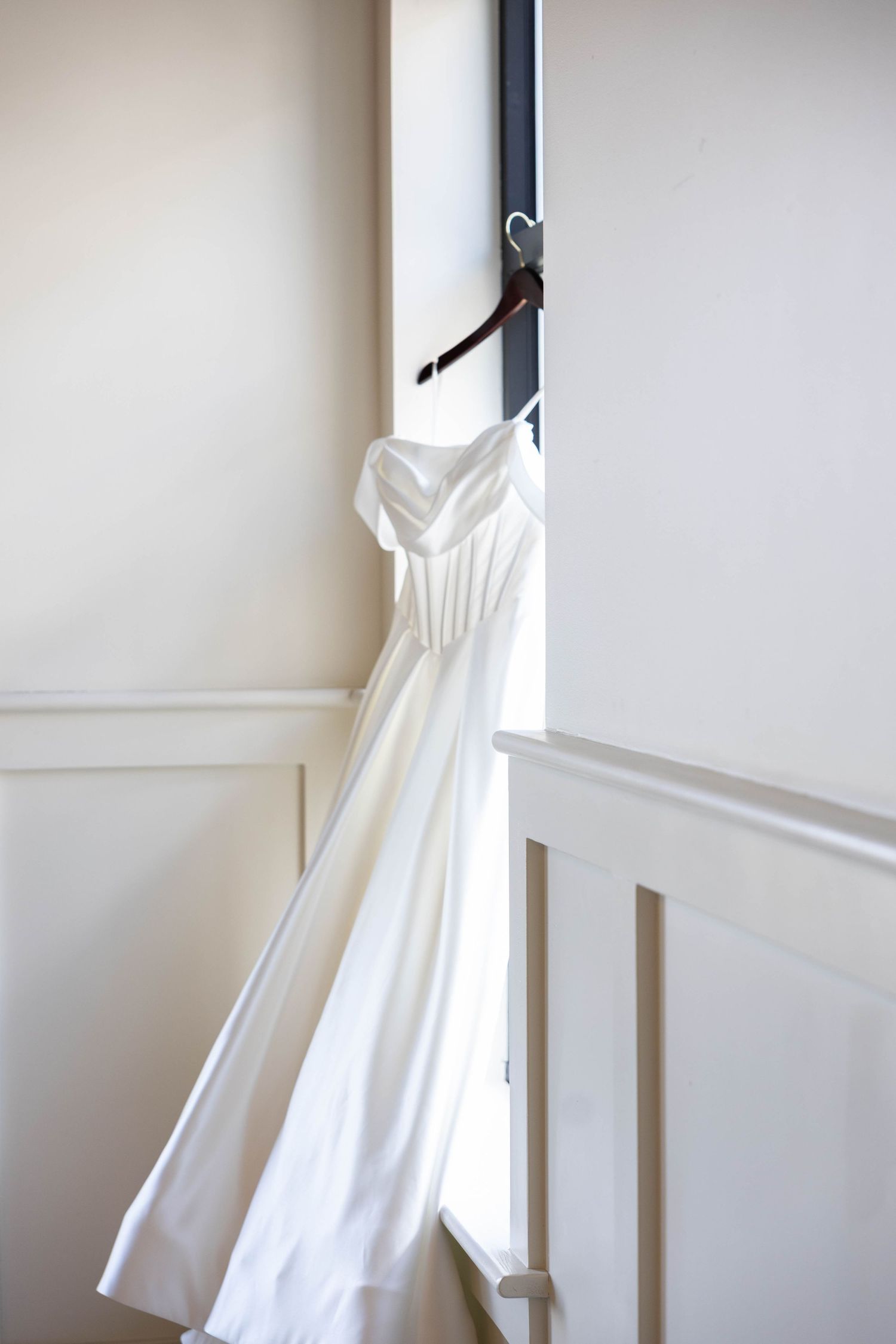Elegant white strapless wedding dress hanging on a wooden hanger by a bright window in a cream-colored room.