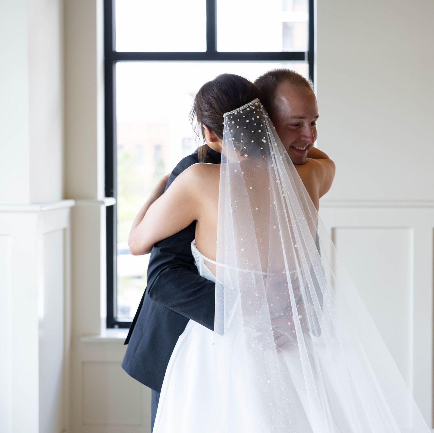 Bride in white gown and pearl veil shares emotional hug with suited guest near window on wedding day.