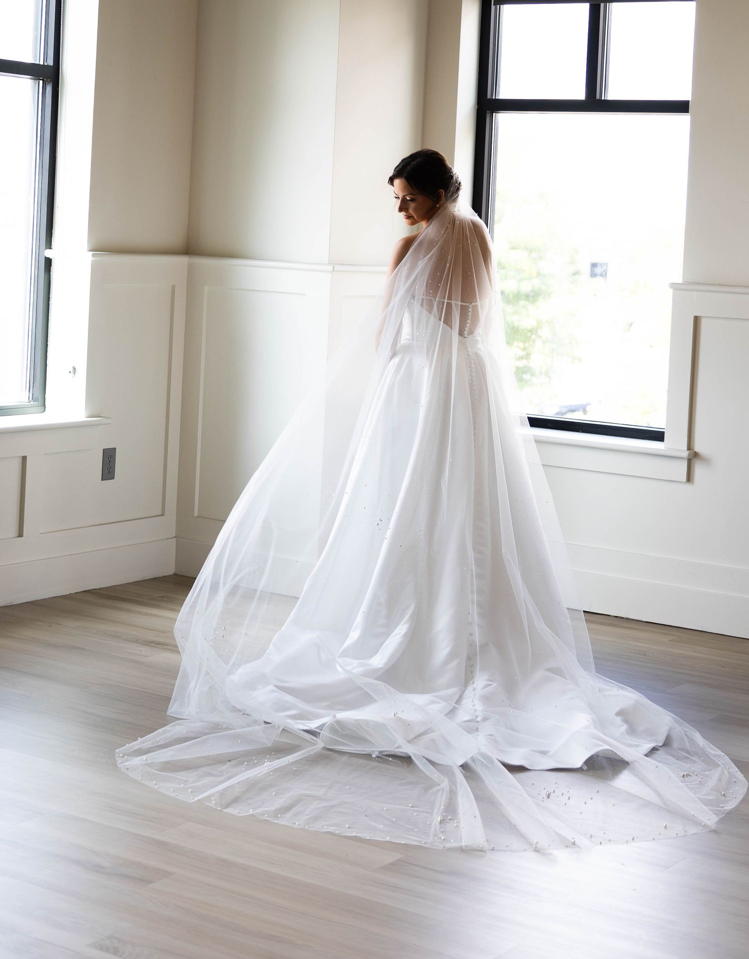 Bride in elegant white wedding gown with long veil and cathedral train standing by bright windows in airy room.