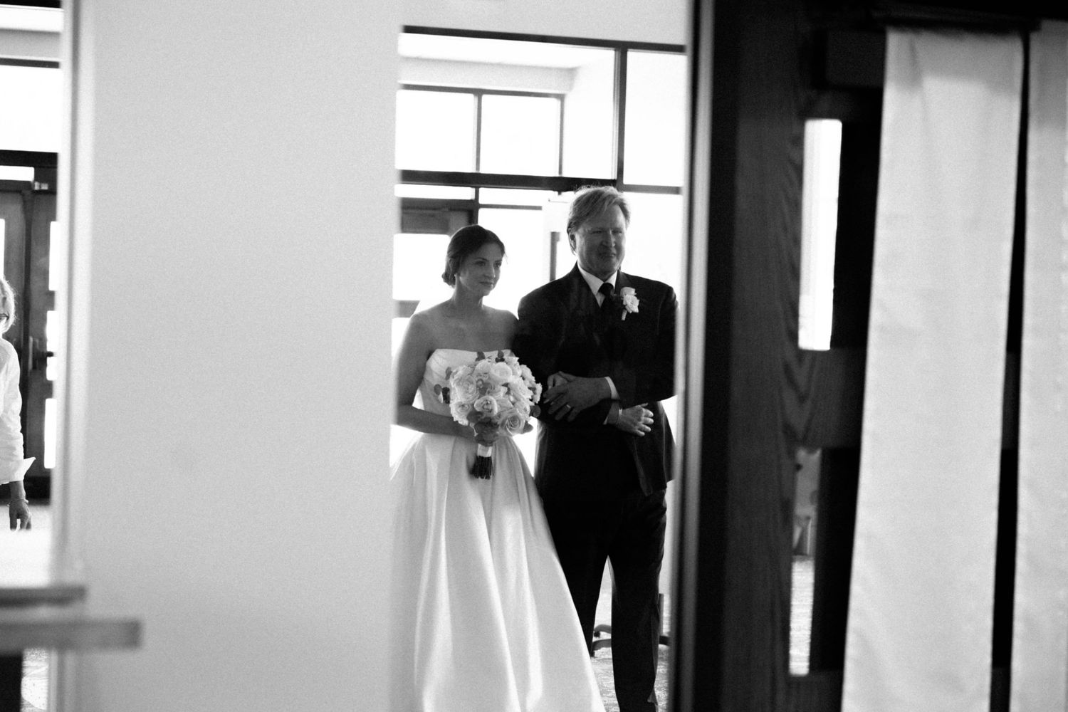 Black and white photo of a bride in a white gown holding a bouquet, walking with an escort through a doorway.