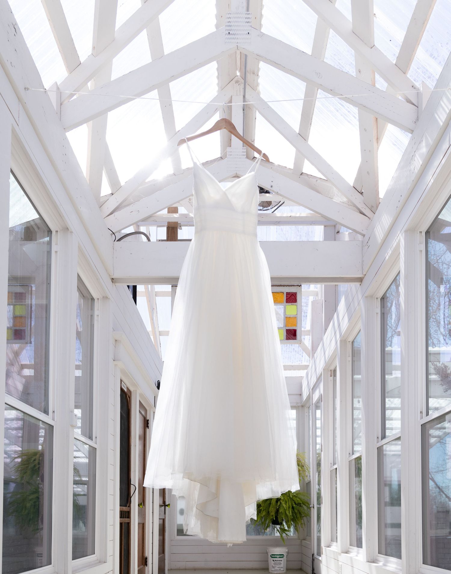 Elegant white wedding dress hanging in sunlit conservatory with glass ceiling and white beams.