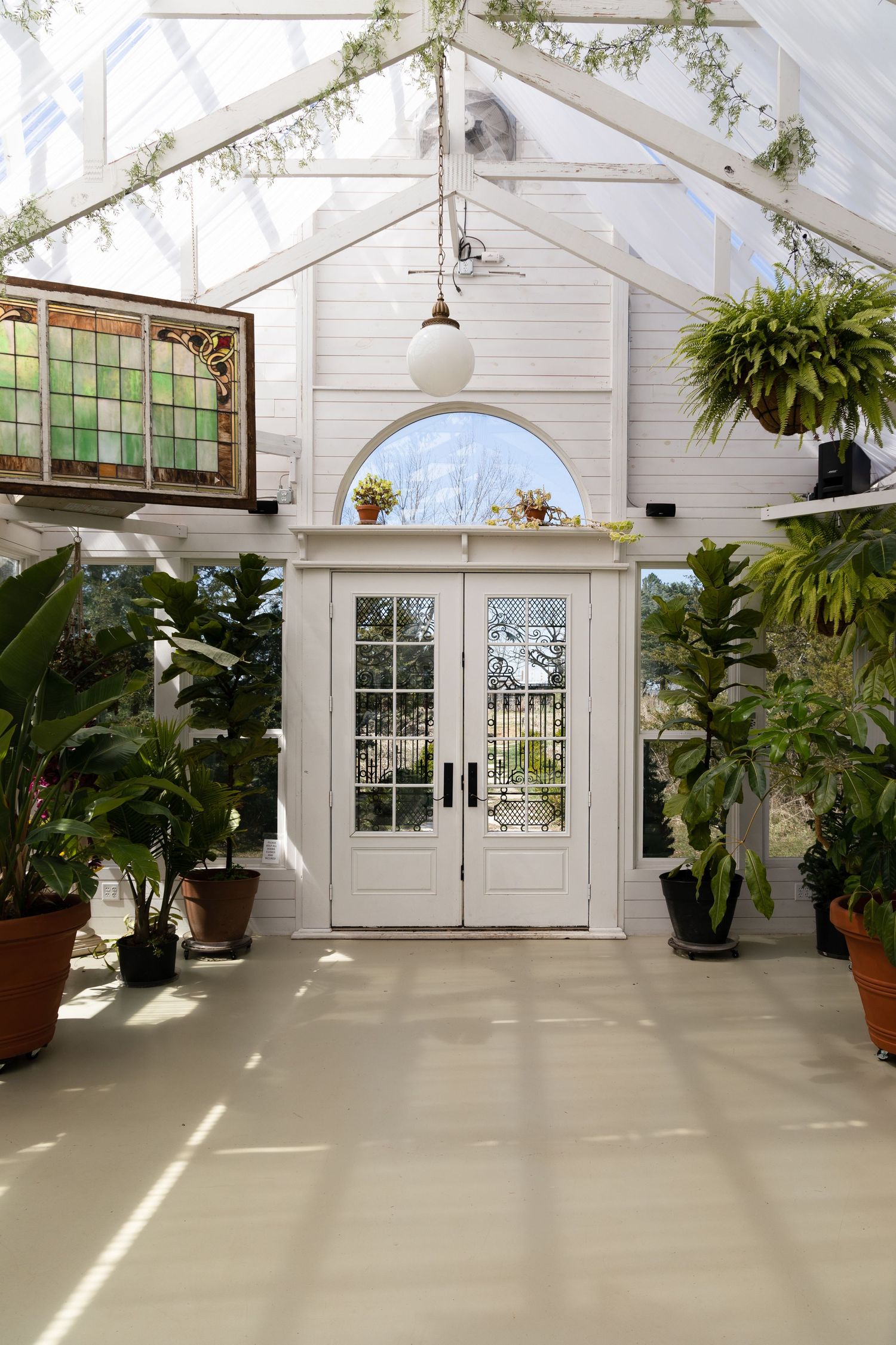 Bright greenhouse interior with white French doors, hanging plants and stained glass window details.