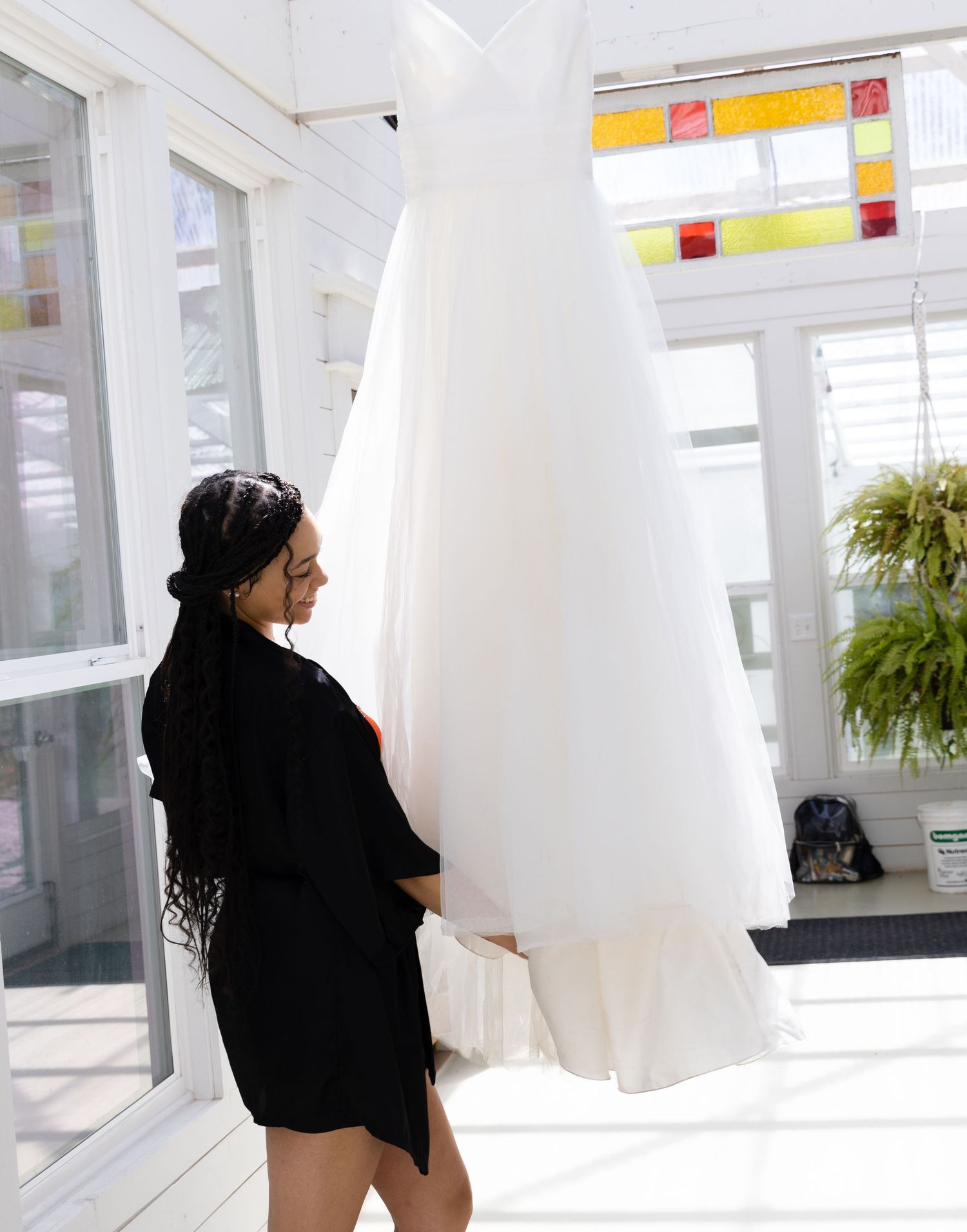 Figure in black robe examining white wedding dress in bright sunlit room.