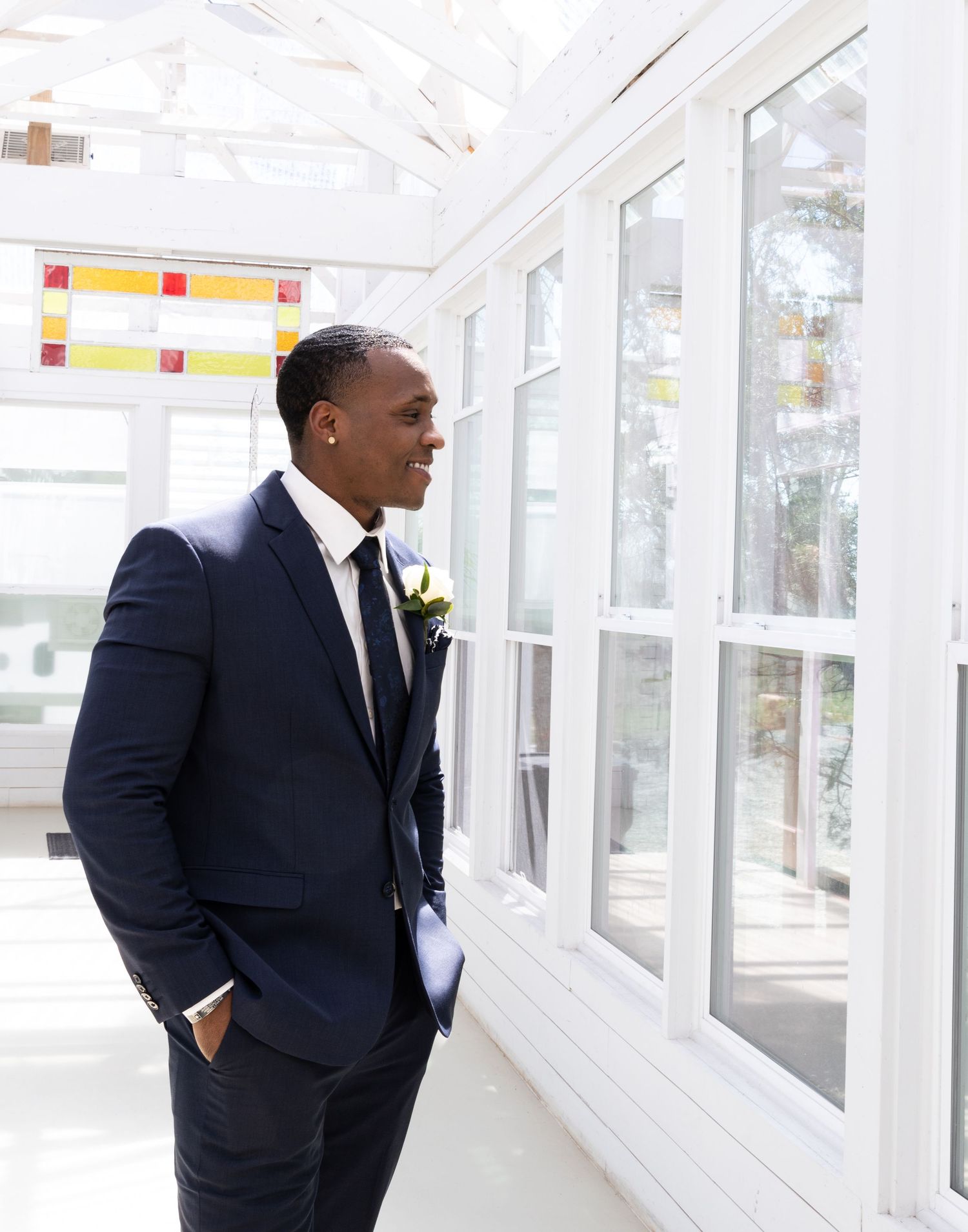 Wedding portrait of groom in dark suit standing by bright windows with boutonniere.
