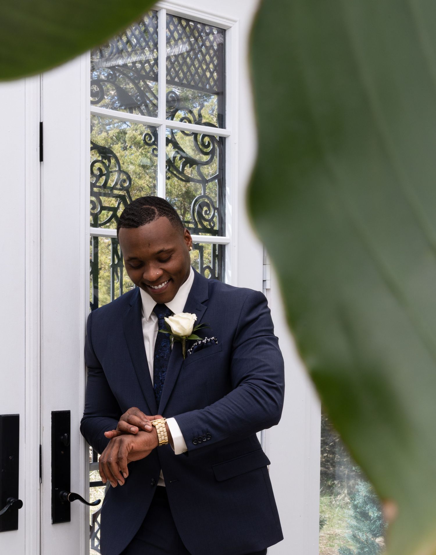 Groom adjusts watch while standing by decorative window in navy suit with white boutonniere.