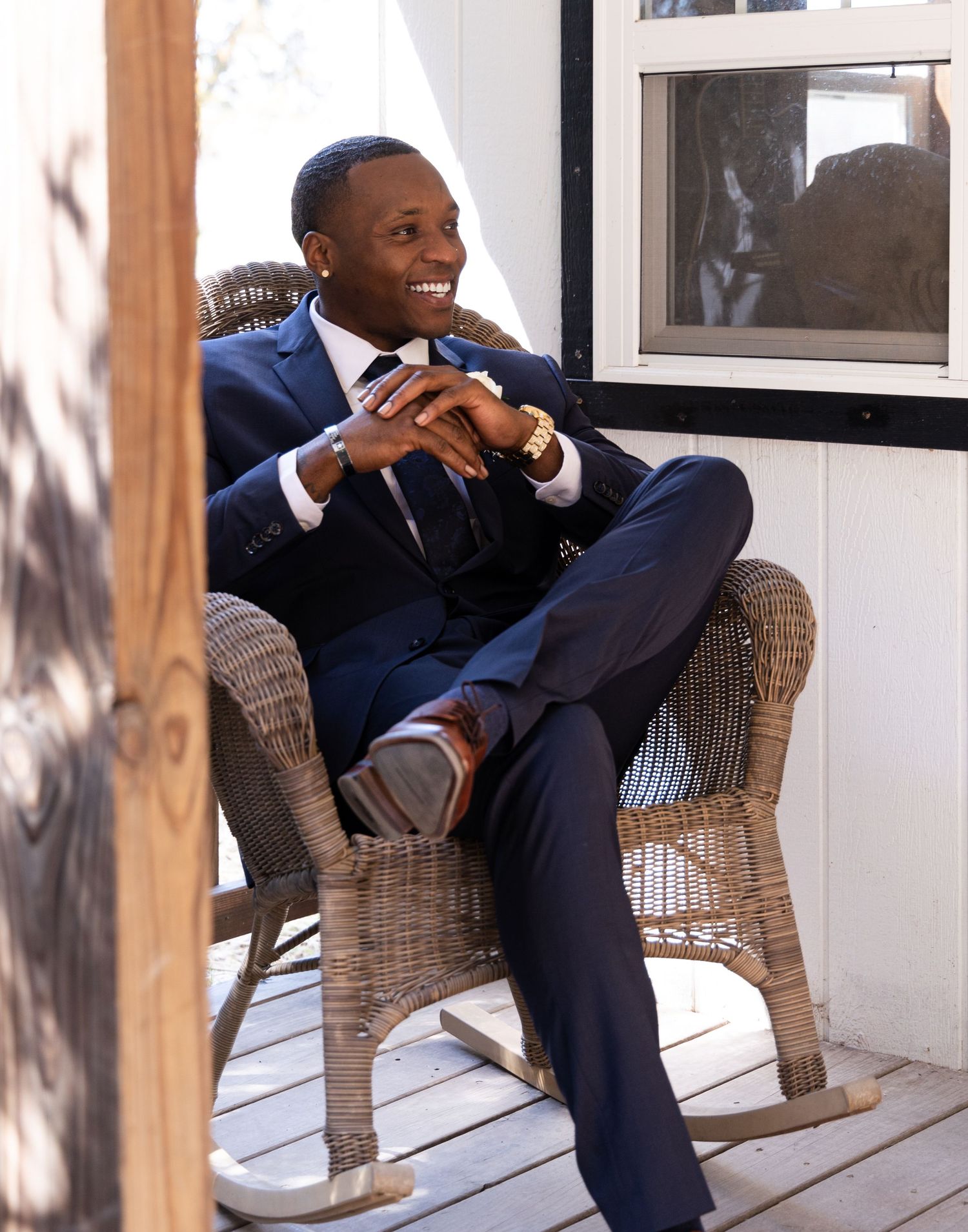 Joyful groom relaxes in wicker chair on porch wearing navy suit.
