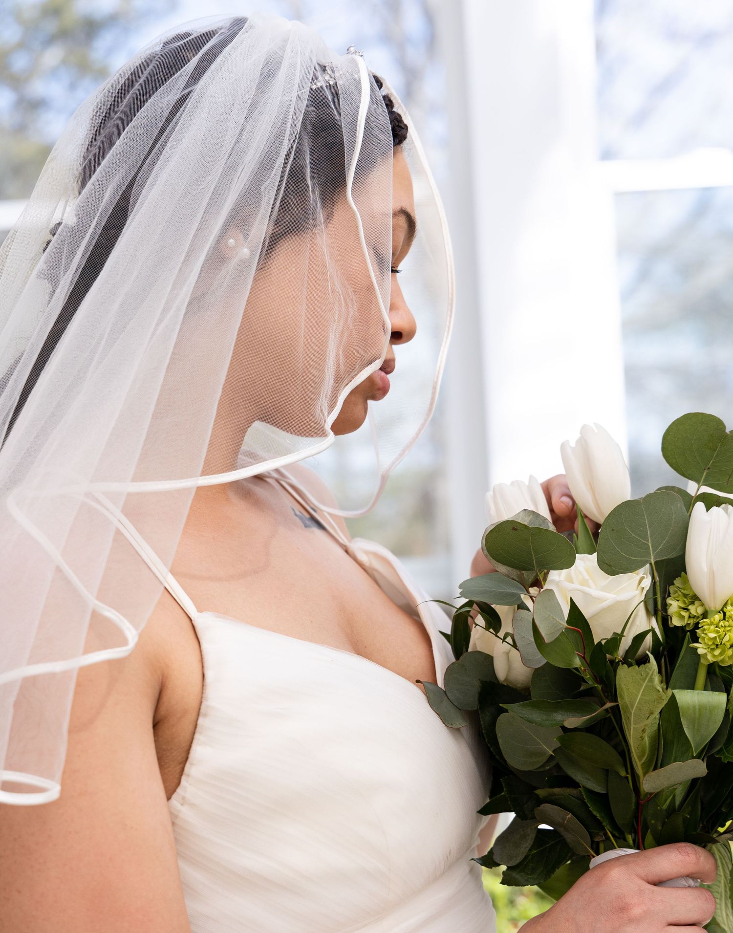 A bride in a white dress with veil holds a bouquet of white roses and eucalyptus against a bright window.