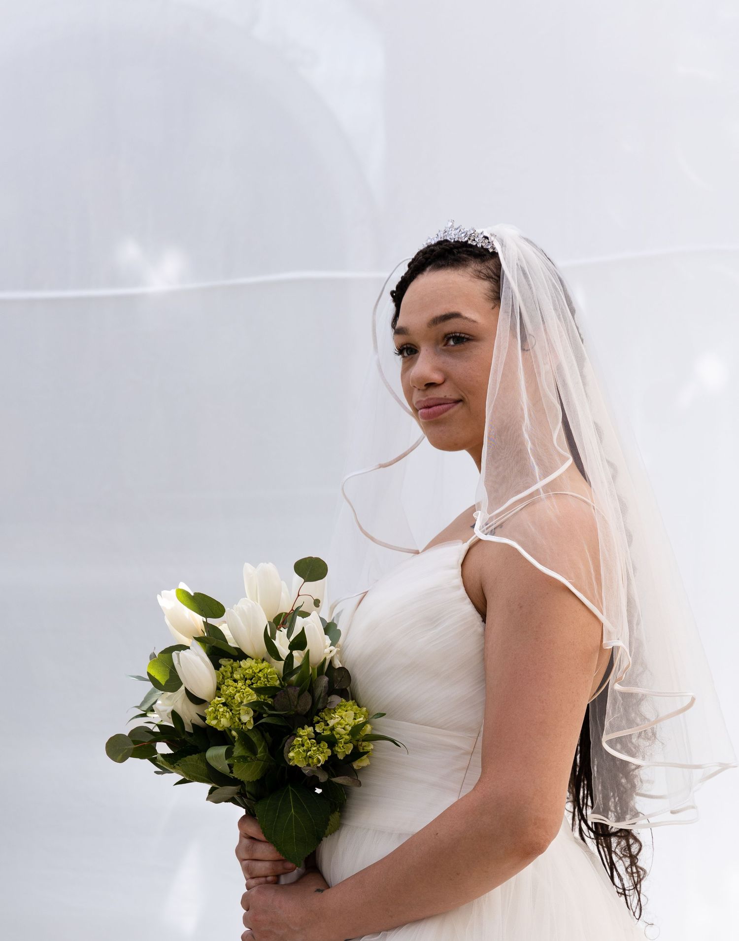 Side view of bride in white dress holding bouquet against light background.