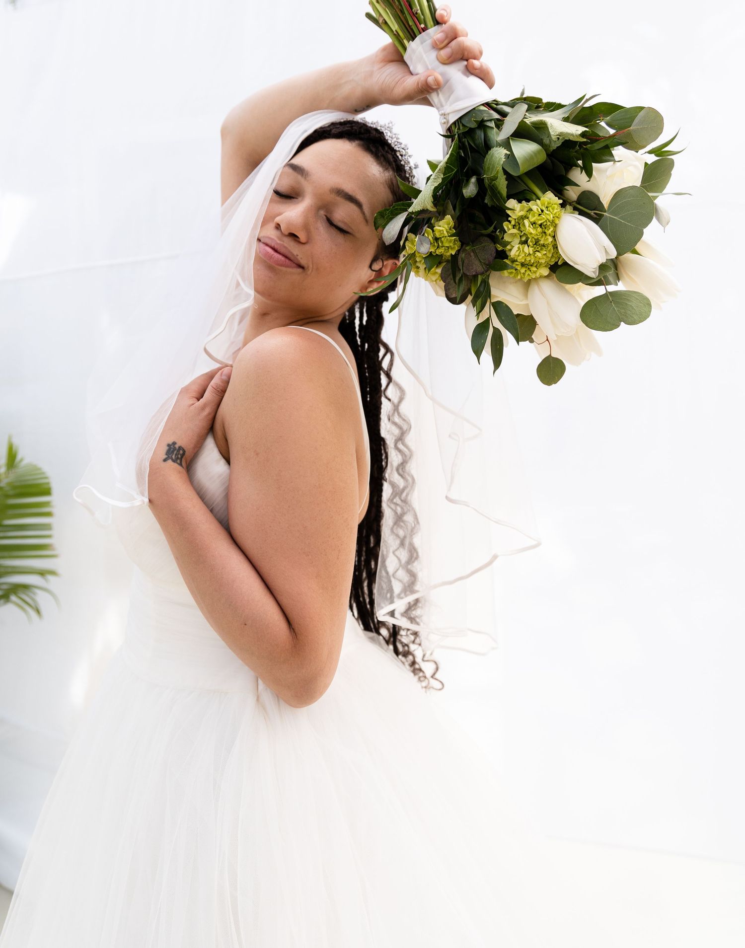 A bride in a white gown holds up her bouquet while posing against a white wall.