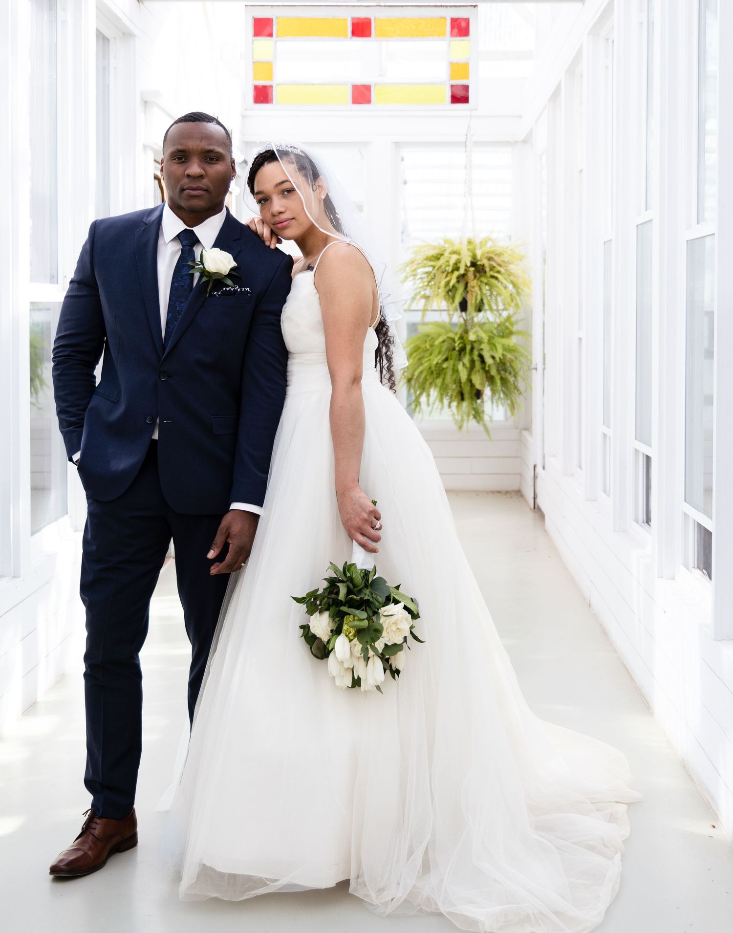 A newlywed couple poses in a bright hallway with hanging plants and colorful accents.