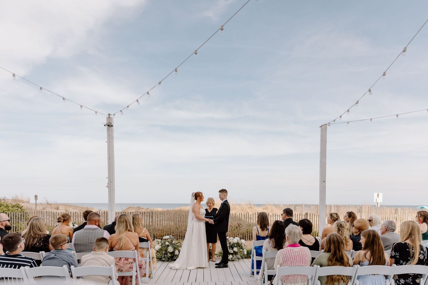 Beach wedding ceremony on wooden deck with string lights and ocean backdrop.