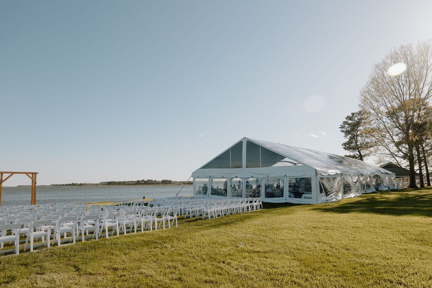 White wedding tent set up on a grassy lawn overlooking water with rows of ceremony chairs.