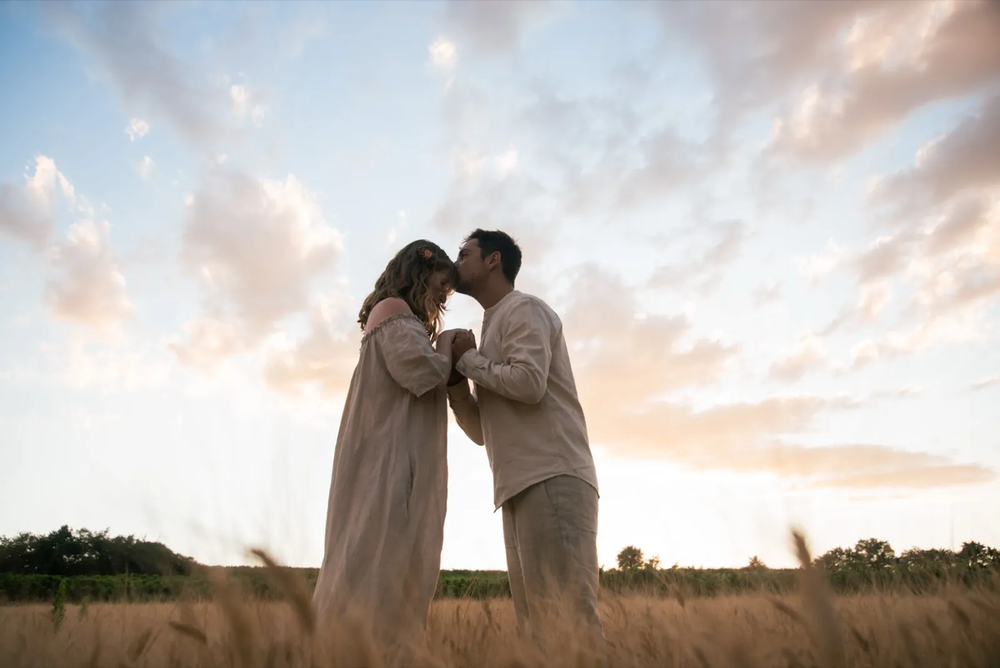 Couple se faisant face dans un champ doré, en train de s’embrasser au crépuscule.