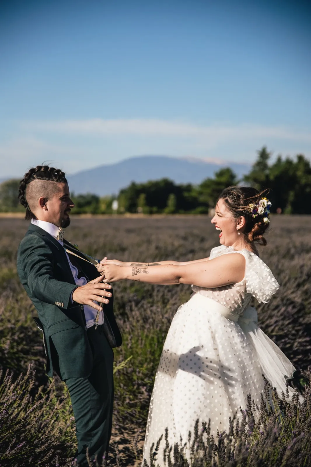 Couple de mariés dans un champ, se tenant par la main avec des montagnes en arrière-plan.