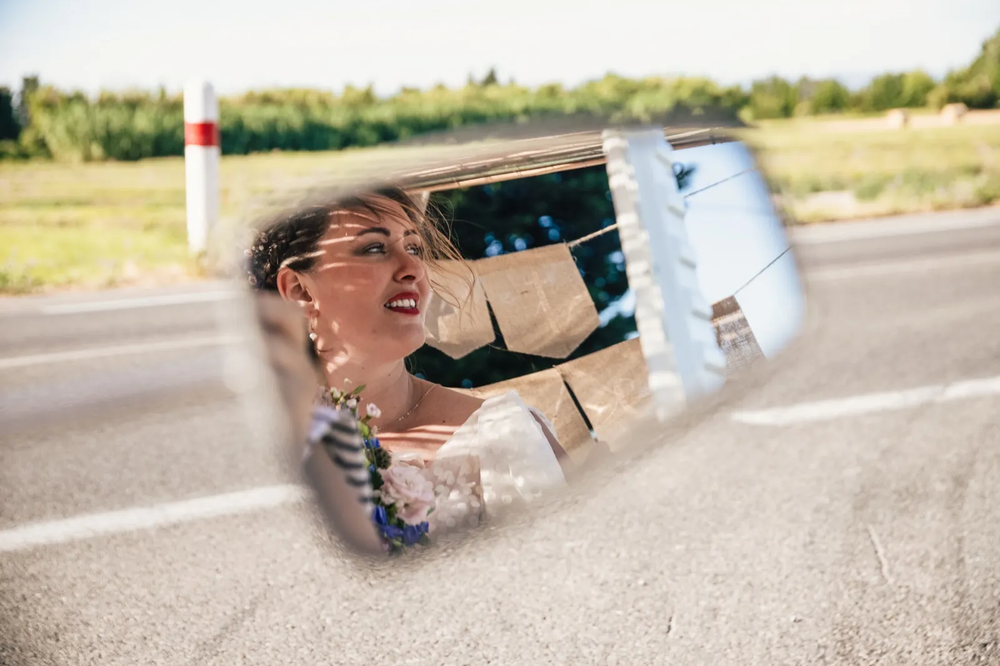 Reflet d’une mariée souriante dans un rétroviseur posé au sol sur une route de campagne.