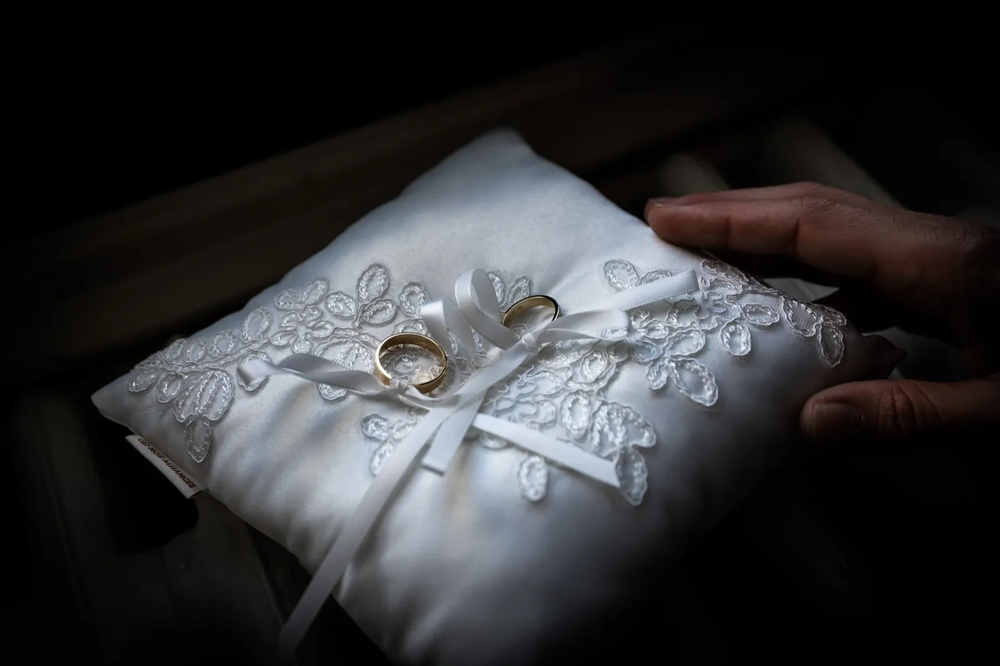 Coussin de mariage en satin blanc avec deux alliances dorées posées au centre, tenu délicatement par une main.