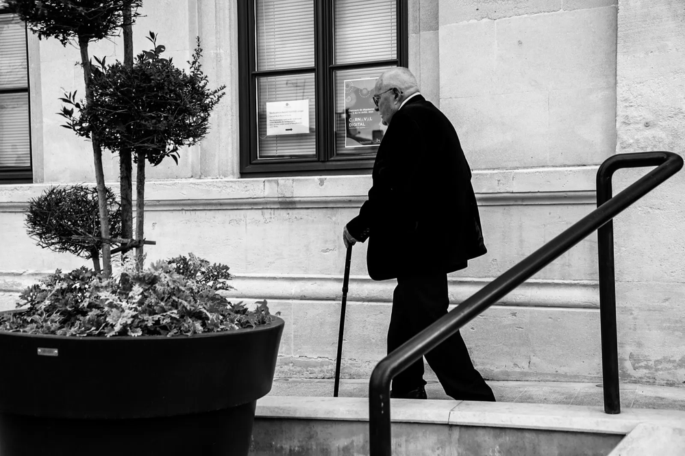 Homme âgé marchant à l’aide d’une canne dans une rue, capté en noir et blanc près d’un bâtiment.