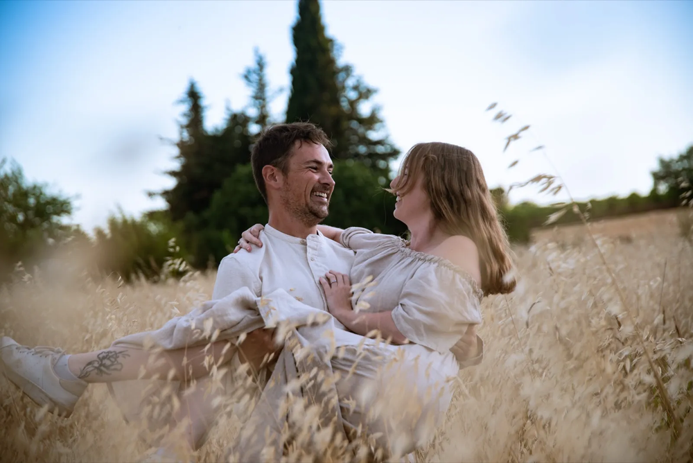 Couple assis dans une prairie, souriant l’un à l’autre au milieu des herbes dorées.