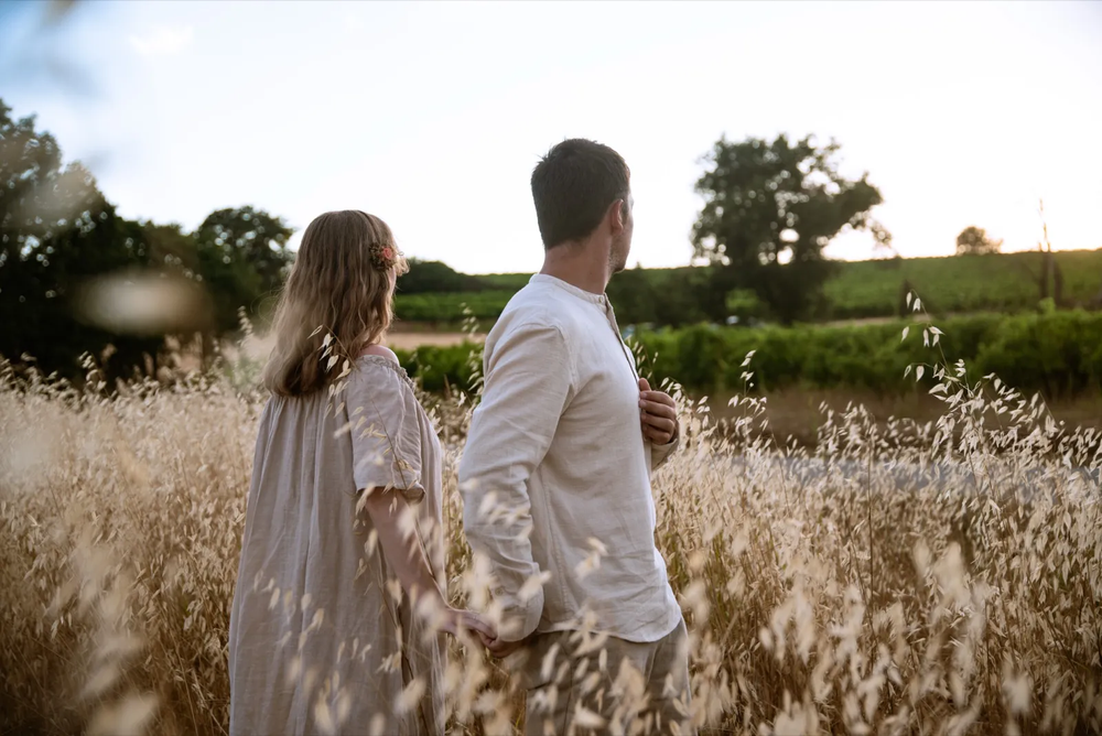 Couple de dos marchant à travers une prairie, entouré de hautes herbes et d’arbres en arrière-plan.