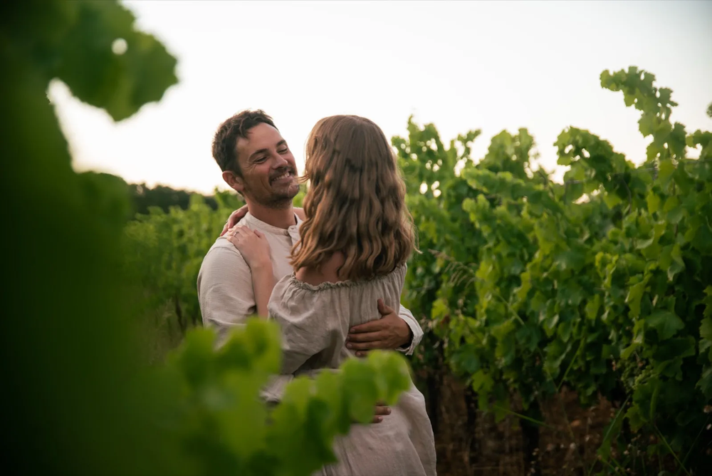 Homme portant tendrement sa compagne dans ses bras entre les rangs de vigne.