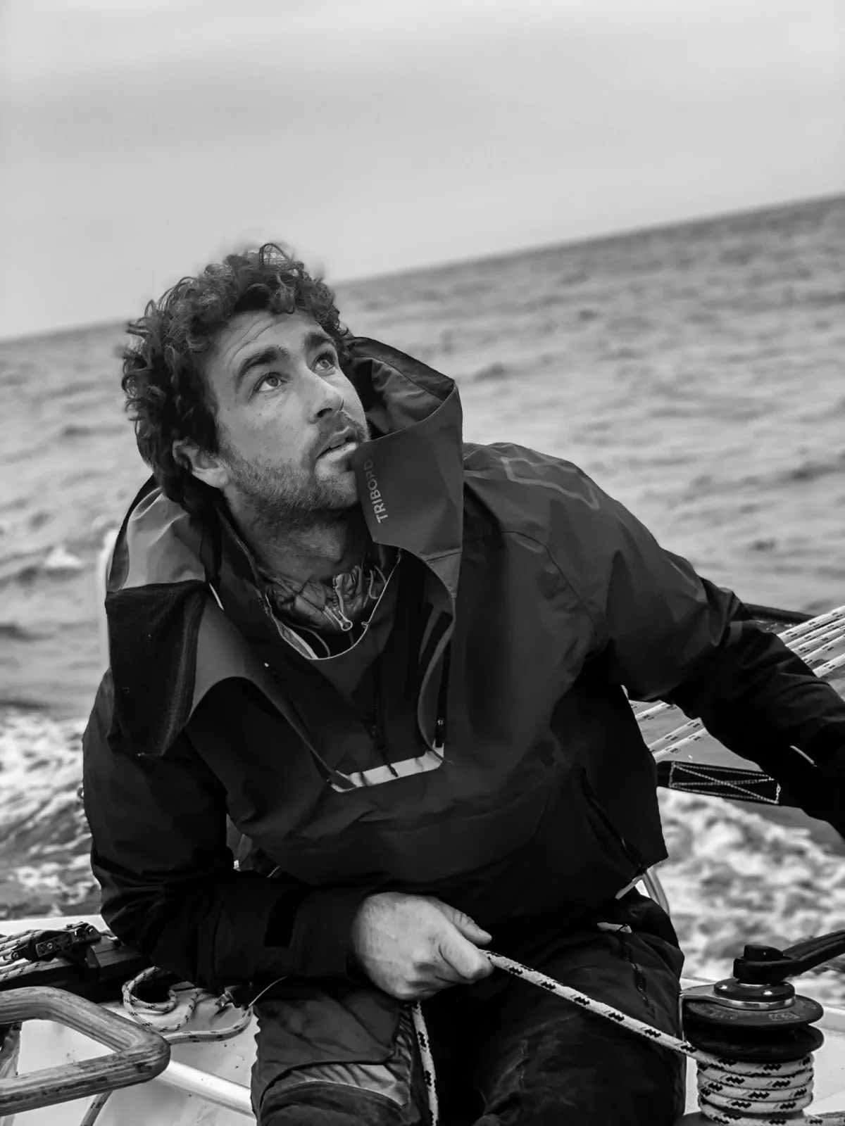 Homme en noir et blanc assis sur un voilier, regardant au loin, avec la mer en arrière-plan.