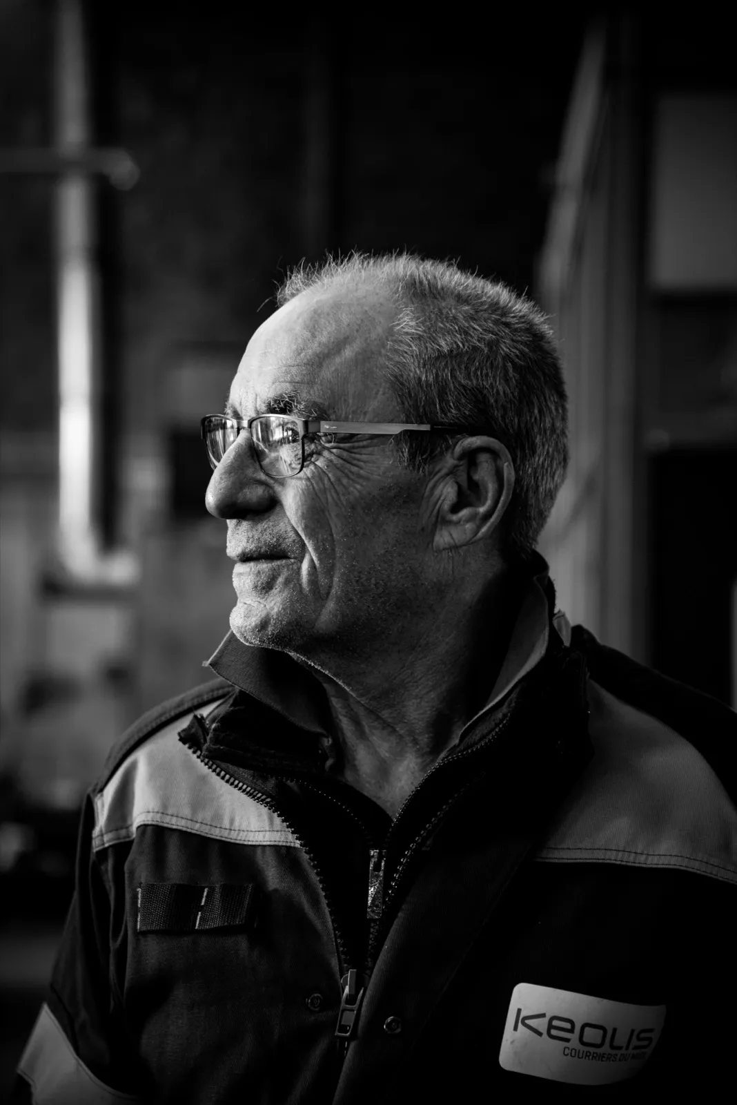 Portrait en noir et blanc d’un homme âgé, en veste de travail, éclairé par une lumière latérale.
