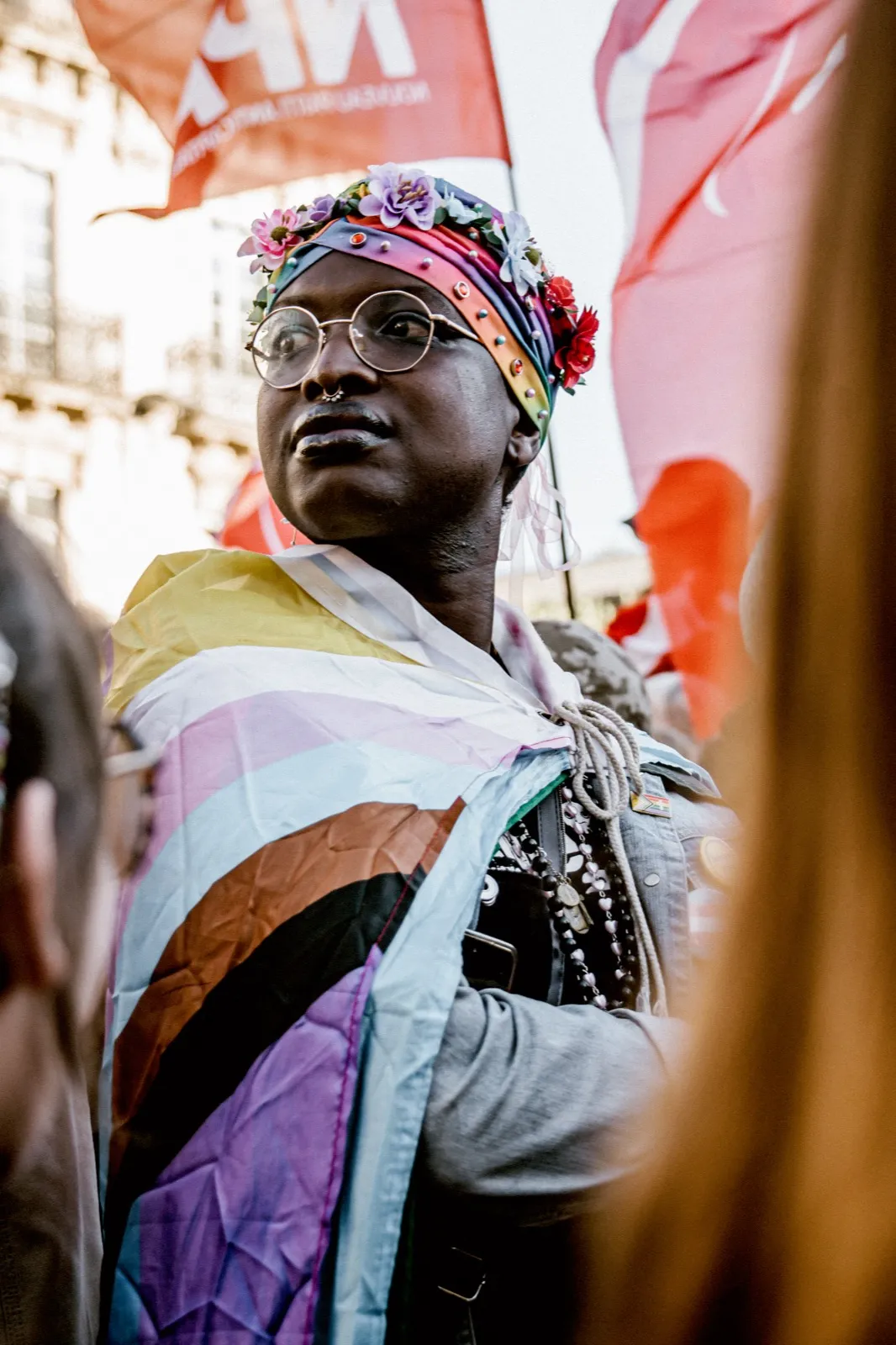 Portrait d’une femme enveloppée dans un drapeau multicolore, regard tourné vers le ciel, lors d’un rassemblement.