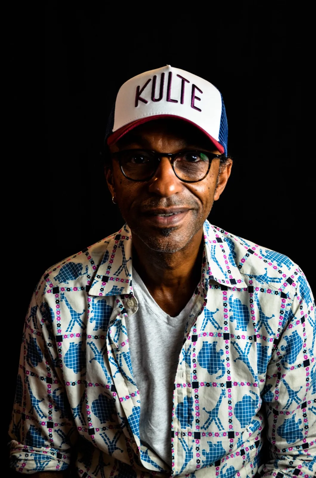 Portrait studio d’un homme vêtu d’une chemise imprimée colorée et d’une casquette “KULTE”, sur fond noir.
