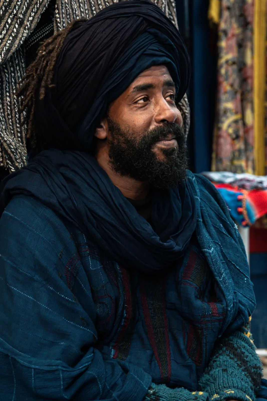 Portrait d’un homme barbu portant un turban et une tenue ample, assis dans une ambiance chaleureuse.
