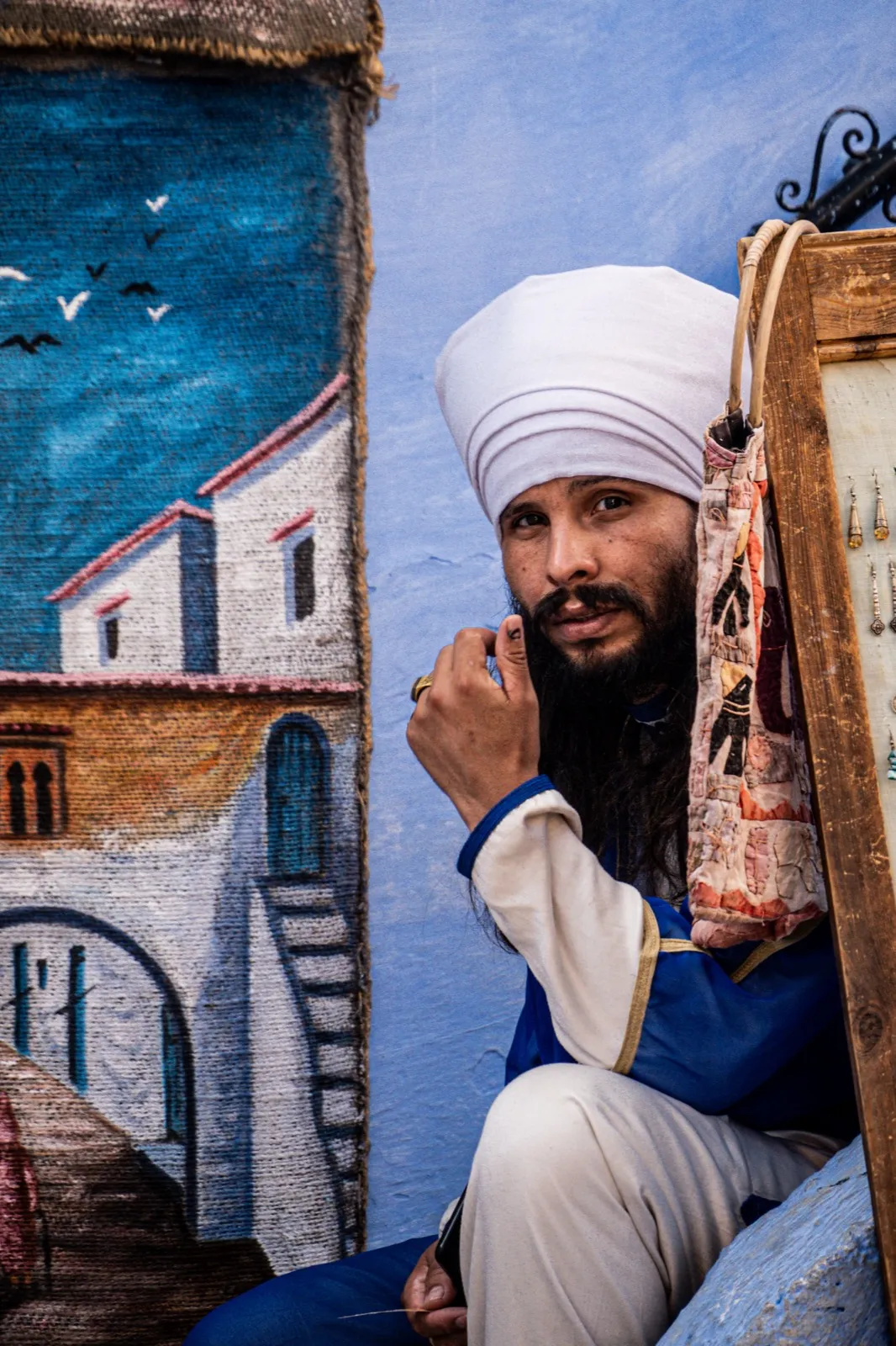 Portrait d’un homme barbu en turban blanc, assis dans une ruelle aux murs peints de fresques colorées.