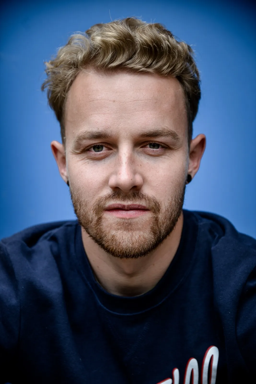 Portrait studio d’un homme aux cheveux courts et barbe naissante, sur fond bleu uni.