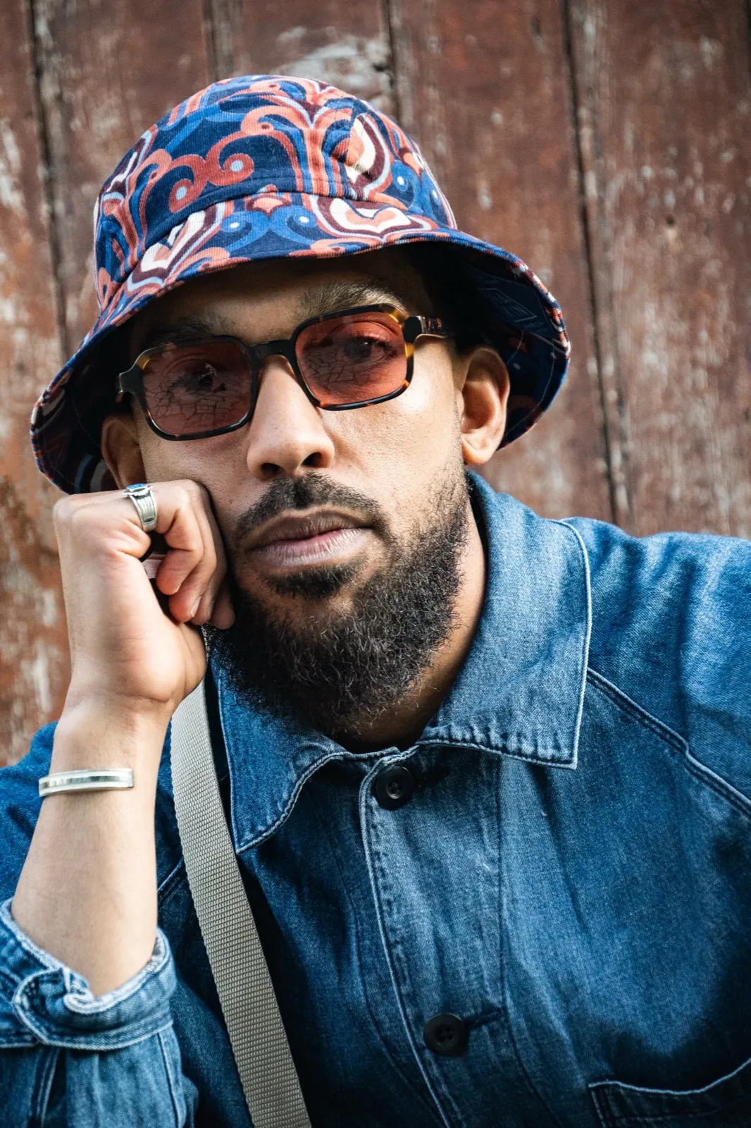 Portrait d’un homme barbu portant un bob fleuri, une chemise en jean et des lunettes de soleil, appuyé contre un mur rouillé.