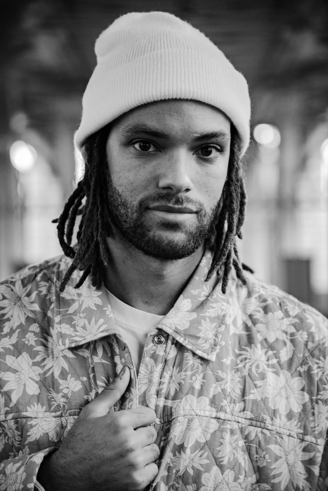 Portrait en noir et blanc d’un homme barbu portant un bonnet et une chemise à motifs, regard intense et main sur le col.