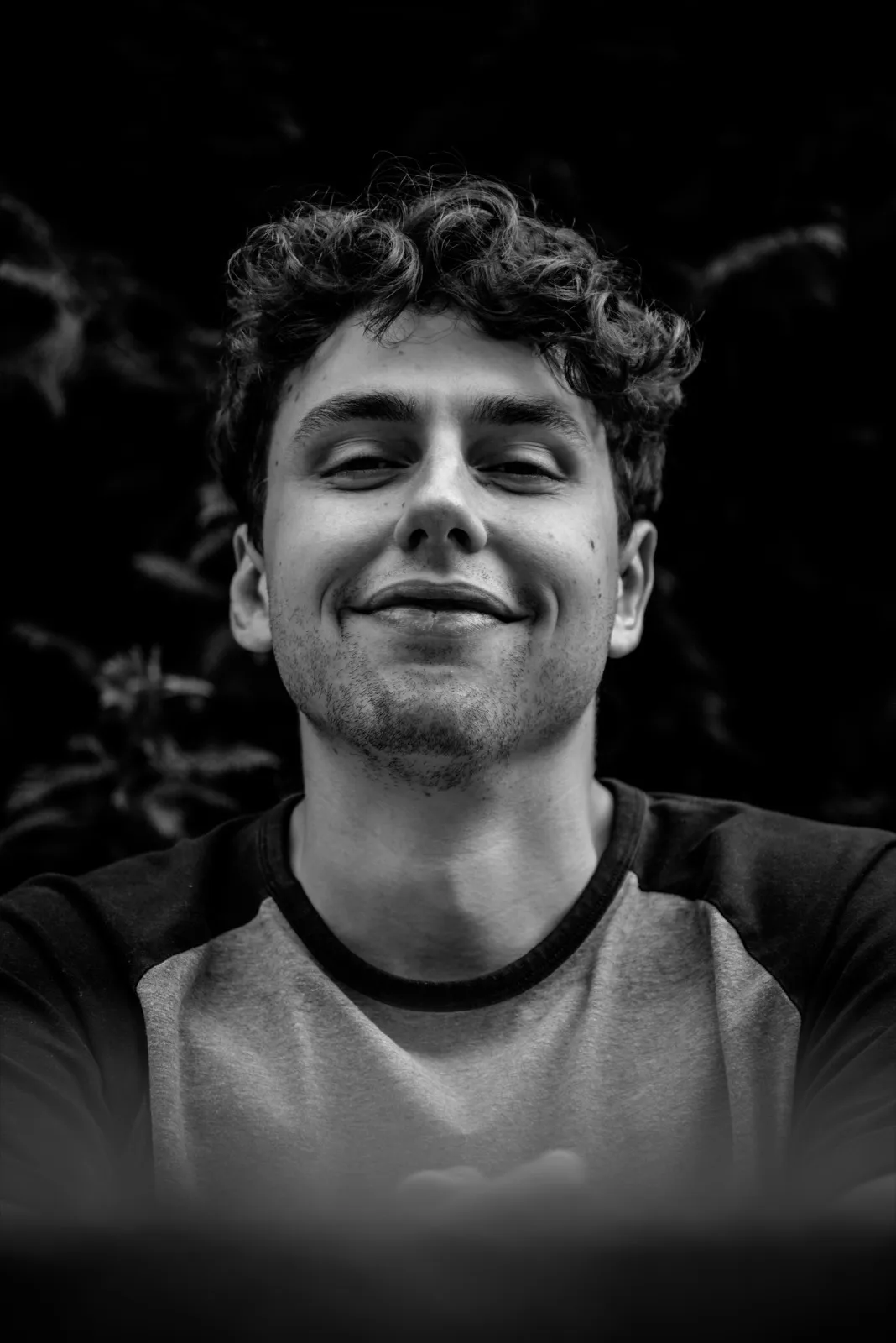 Portrait d’un jeune homme souriant sous une lumière tamisée, fond sombre et feuillage flou.