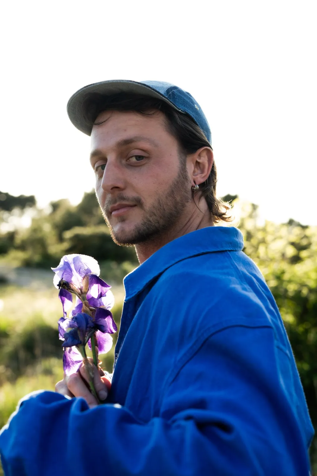 Portrait d’un homme en bleu avec une casquette, tenant un bouquet de fleurs violettes en extérieur.