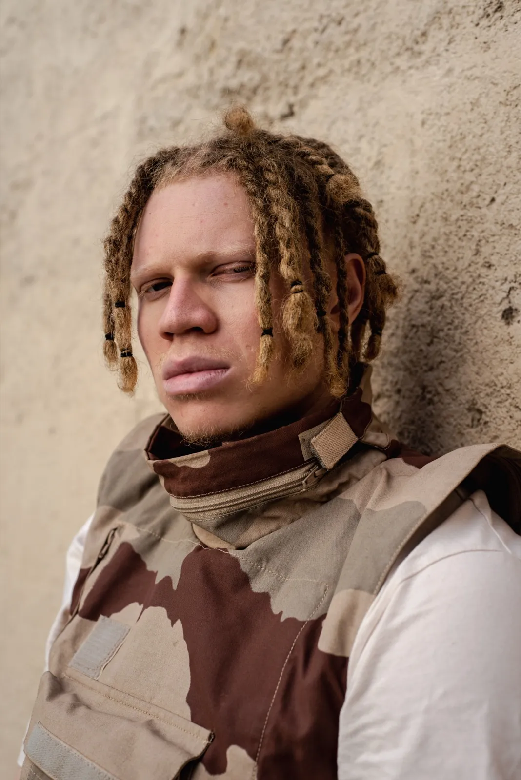 Jeune homme aux dreadlocks, portant un débardeur camouflage, posé devant un mur beige.