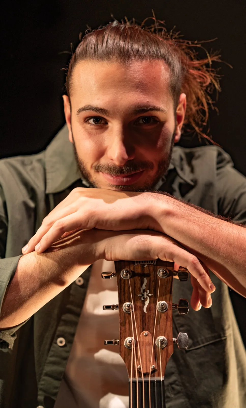 Portrait serré d’un homme aux cheveux tirés en arrière, posant les bras croisés sur une guitare, dans une lumière chaude.