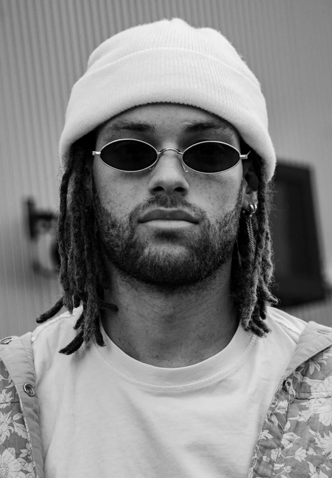 Homme portant un bonnet blanc, des lunettes de soleil et une coupe dreadlocks, photographié en noir et blanc.