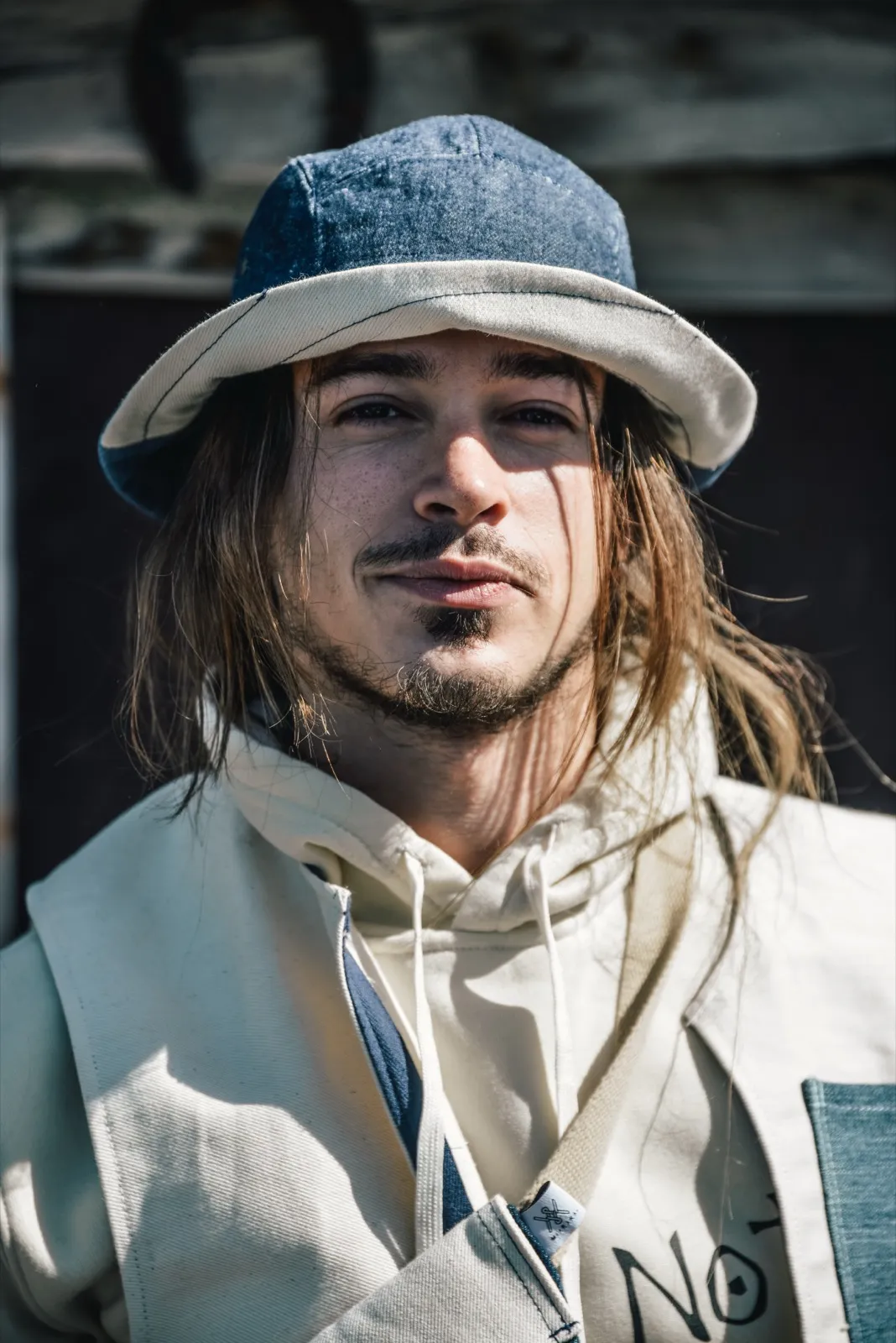 Homme aux cheveux longs et bouclés, portant un bob bleu clair et un sweat, en extérieur.