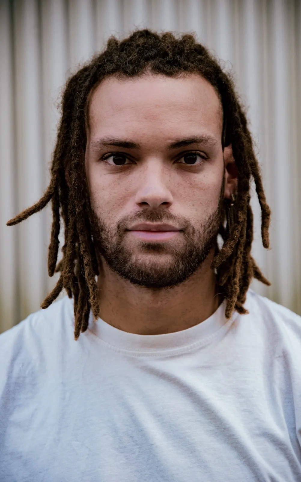 Portrait d’un homme aux dreadlocks et t-shirt blanc, posé devant une surface en métal ondulé.