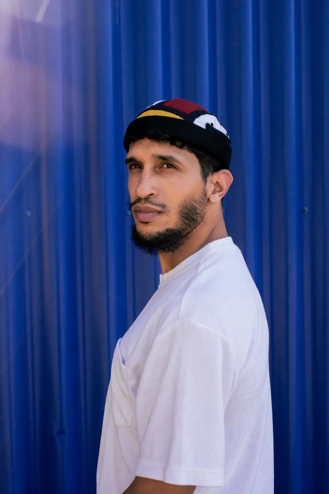 Jeune homme en t-shirt blanc et miki, de profil, devant une paroi bleu métallique.