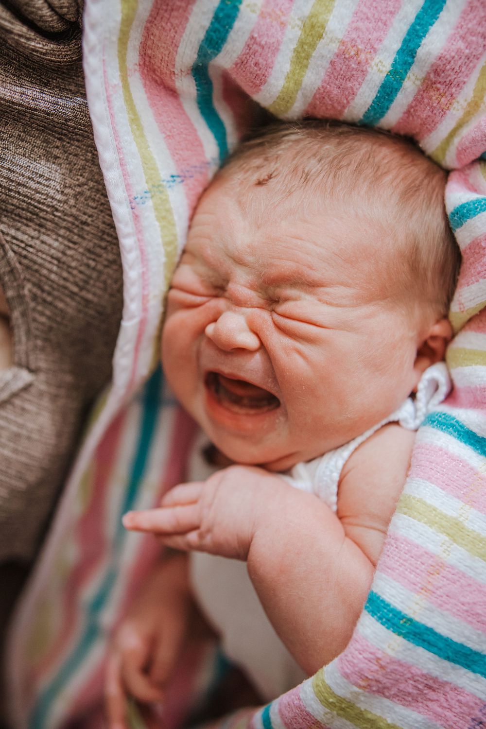 Crying newborn baby wrapped in colorful striped blanket with pink, teal and yellow stripes.