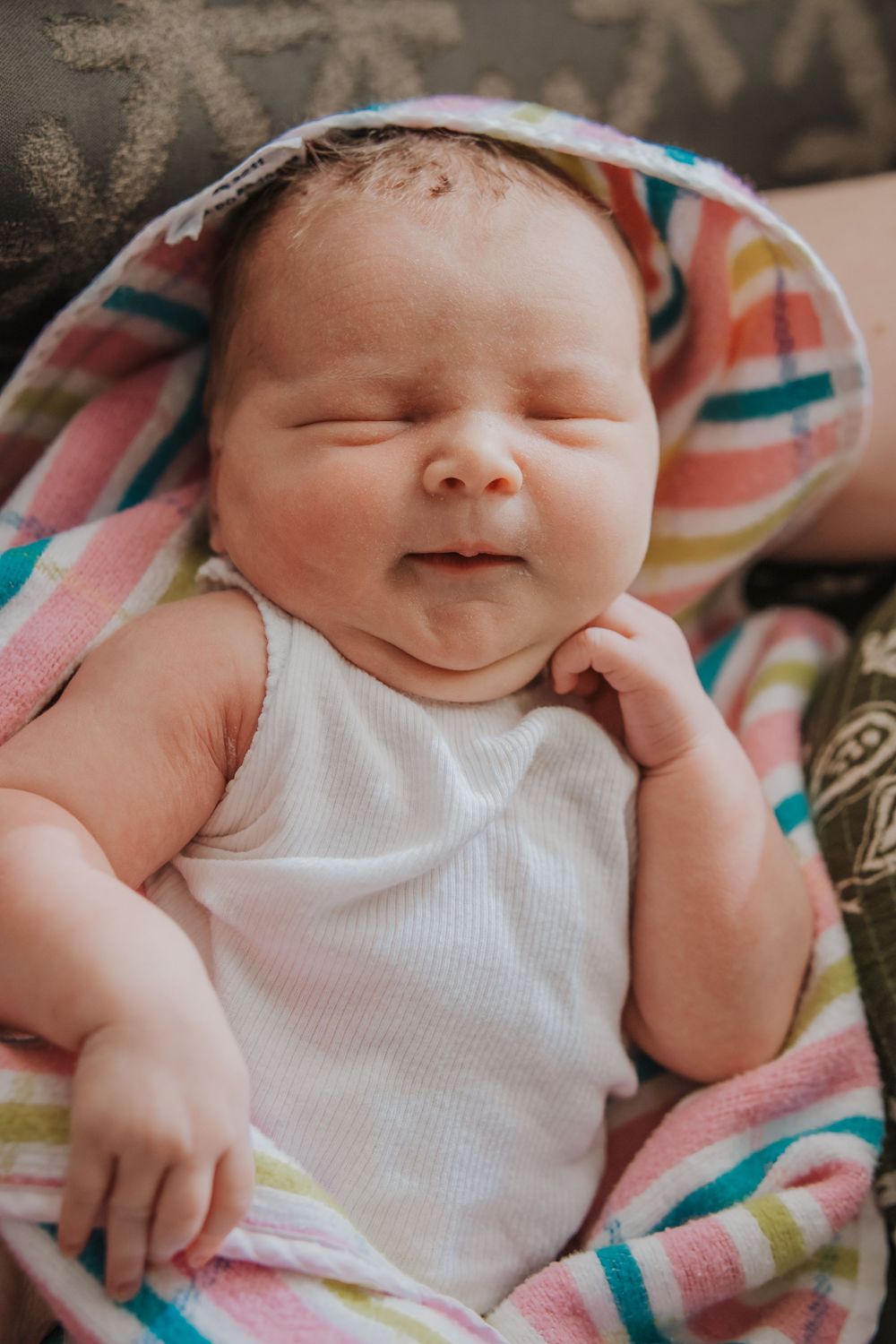 A content newborn baby wearing a white onesie smiles while wrapped in a colorful striped blanket.