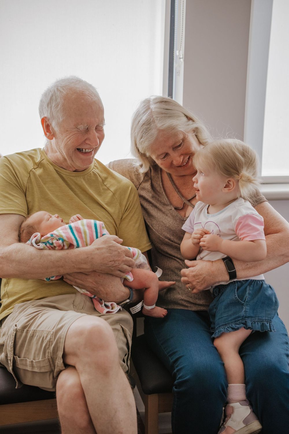Three generations share laughter while bonding with a newborn on a window bench.