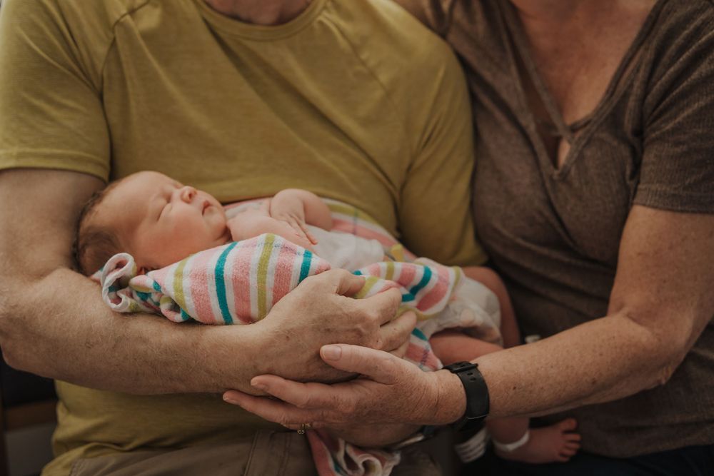 Two people hold a sleeping newborn baby wearing a colorful striped onesie while sitting together.
