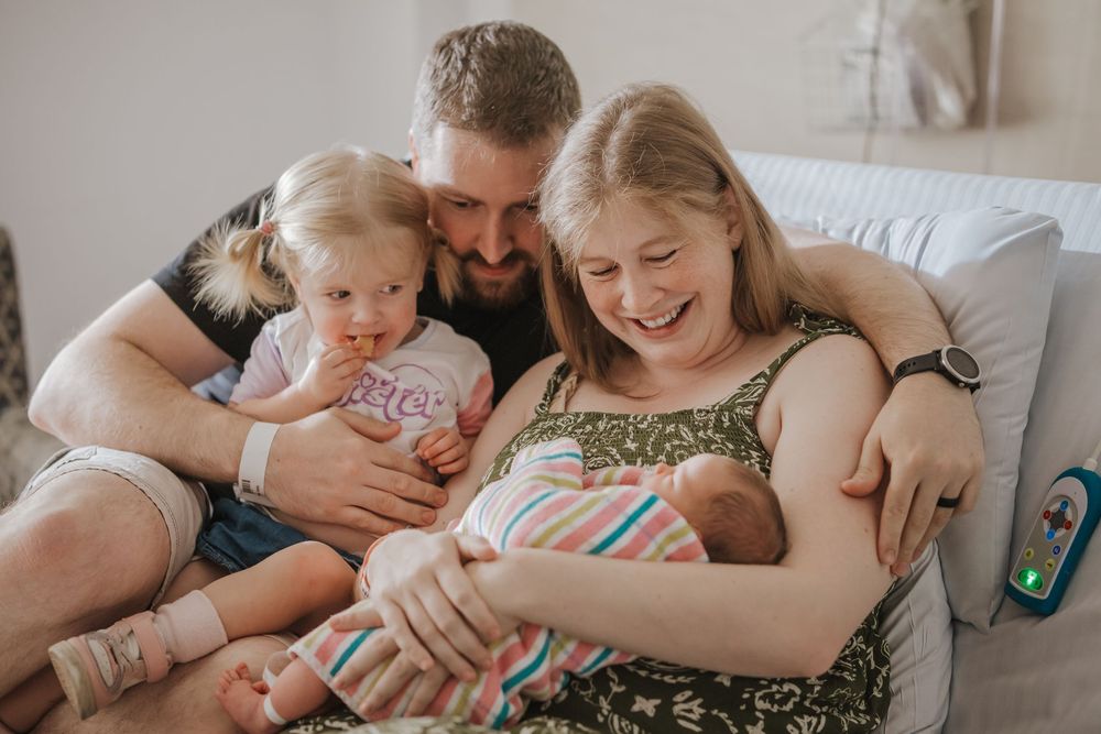 A family cuddles together on a couch while bottle feeding a newborn baby at home.